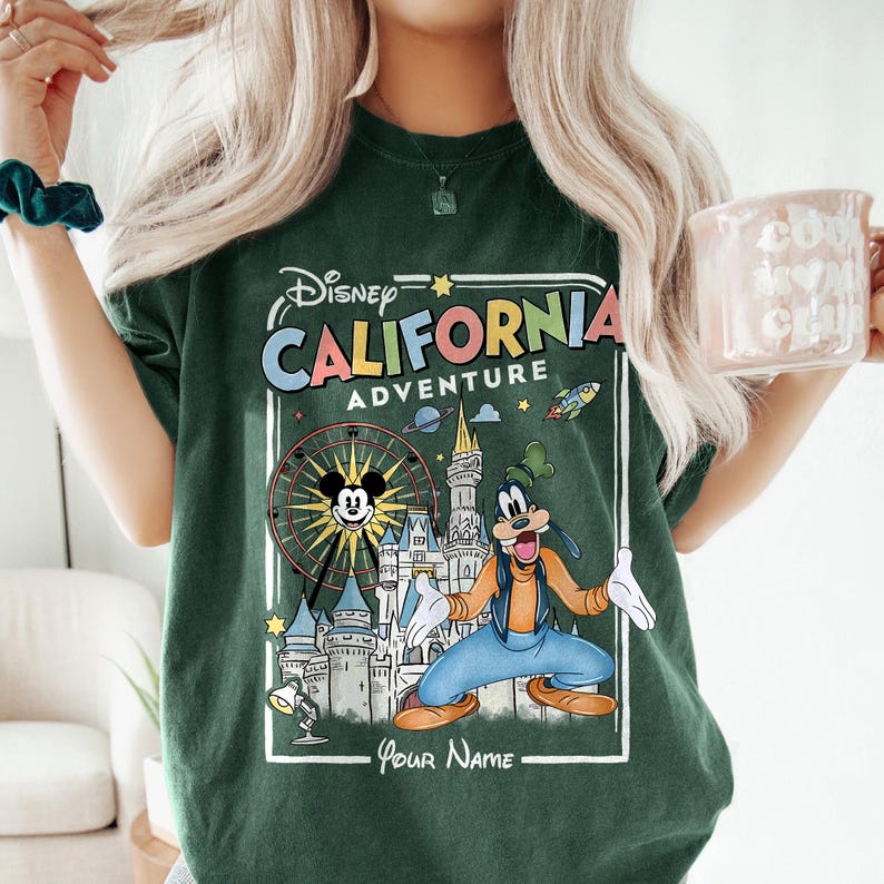 Puede incluir: Camiseta verde oscuro con un gr&aacute;fico colorido de Disney's California Adventure. El dise&ntilde;o incluye a Mickey Mouse, Goofy, una noria y el texto "California Adventure". La camiseta es de corte holgado.
