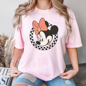 以下が含まれることがあります： ミニーマウスのグラフィックが特徴のライトピンクのTシャツ。グラフィックは、桃色のリボンが付いたミニーの顔を、白黒のチェック柄の円で囲んでいます。このシャツは、カジュアルな半袖スタイルです。