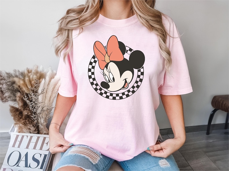 以下が含まれることがあります： ミニーマウスのグラフィックが特徴のライトピンクのTシャツ。グラフィックは、桃色のリボンが付いたミニーの顔を、白黒のチェック柄の円で囲んでいます。このシャツは、カジュアルな半袖スタイルです。