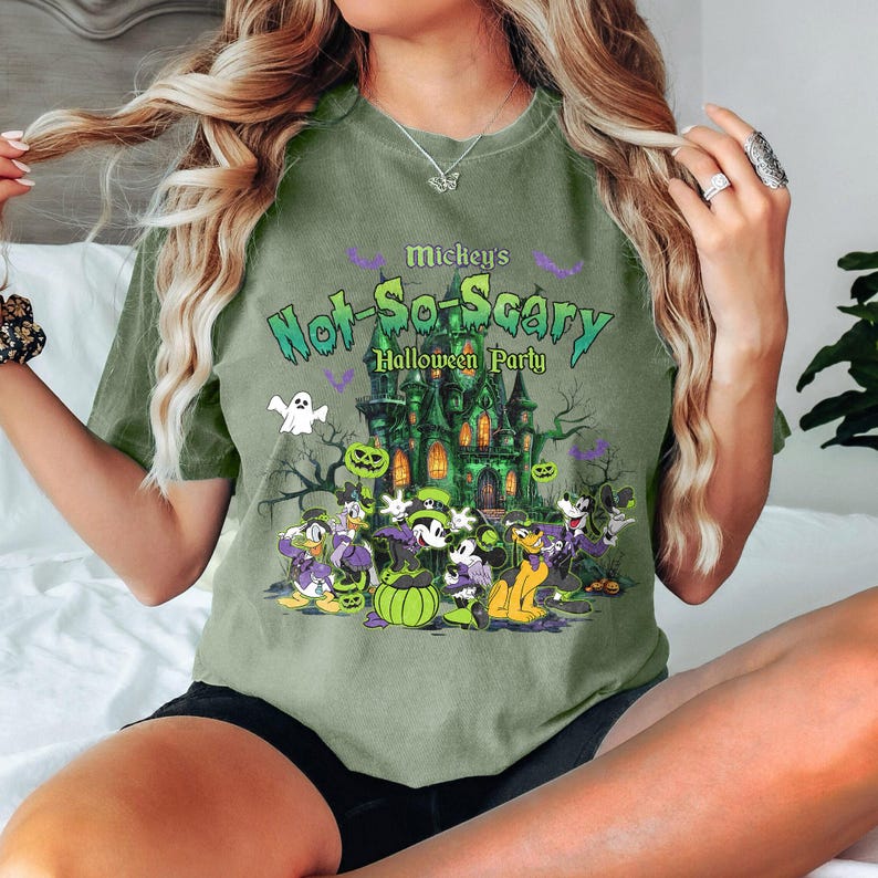 May include: Olive green t-shirt featuring a graphic for Mickey's Not-So-Scary Halloween Party. The design includes a haunted house, Disney characters, pumpkins, and the text "Mickey's Not-So-Scary Halloween Party".