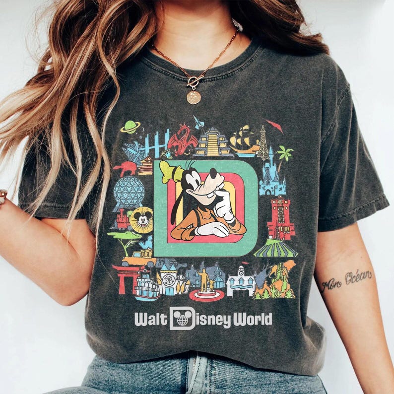 Puede incluir: Camiseta gris oscuro con un gr&aacute;fico colorido de Goofy dentro de una gran "D", rodeado de monumentos de Disney World. Las palabras "Walt Disney World" est&aacute;n impresas debajo en blanco.