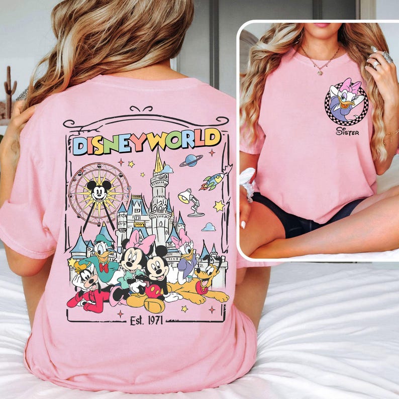 Op de afbeelding: Lichtroze T-shirt met een Disney World-afbeelding op de achterkant, met Mickey Mouse, Minnie Mouse, Donald Duck, Goofy en Pluto. De voorkant heeft een kleine Daisy Duck-afbeelding en het woord "Sister".