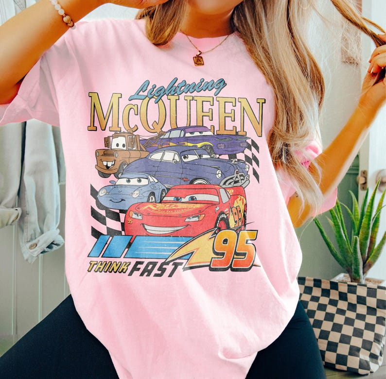 May include: Pink t-shirt with a graphic print of Lightning McQueen and other Cars characters. The text "Lightning McQueen" and "Think Fast" are printed on the shirt.