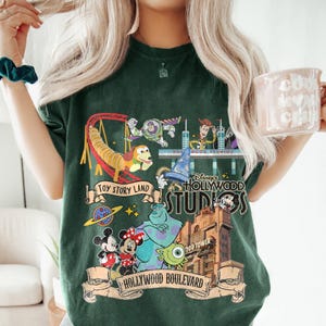 May include: Dark green t-shirt featuring a colorful graphic of Disney's Hollywood Studios. The design includes characters from Toy Story, Mickey and Minnie Mouse, and the Hollywood Tower Hotel. The words "Toy Story Land" and "Hollywood Boulevard" are also visible.