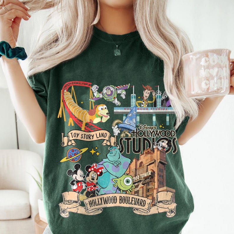 May include: Dark green t-shirt featuring a colorful graphic of Disney's Hollywood Studios. The design includes characters from Toy Story, Mickey and Minnie Mouse, and the Hollywood Tower Hotel. The words "Toy Story Land" and "Hollywood Boulevard" are also visible.