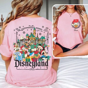 May include: Pink t-shirt featuring a Disneyland graphic with Mickey Mouse, Donald Duck, and other Disney characters in front of a castle. The shirt also has a small Daisy Duck graphic with the word "Sister".