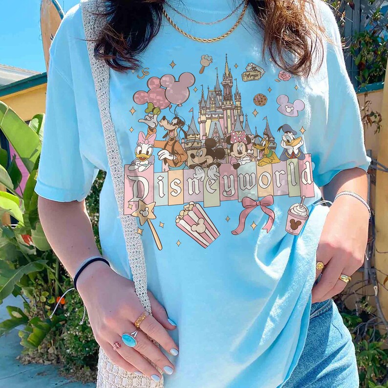 May include: Light blue t-shirt featuring a colorful Disney World graphic. The design includes Mickey Mouse, Minnie Mouse, Donald Duck, and other characters, along with a castle, popcorn, and the text "Disneyworld".