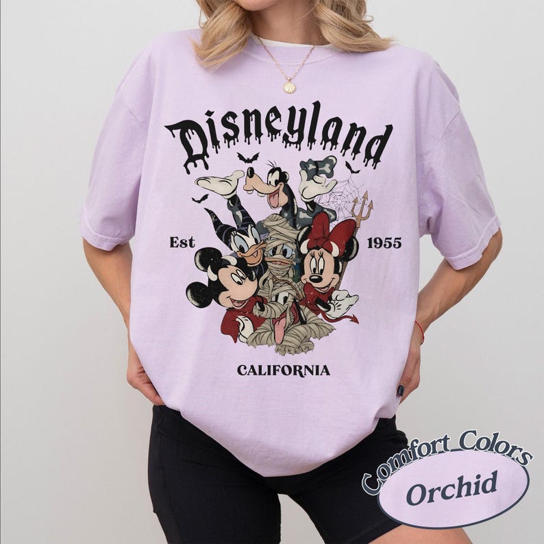 May include: A light purple t-shirt featuring a graphic print with the text Disneyland Est 1955 California. The design includes cartoon characters dressed in Halloween costumes, including a mummy, a devil, and a vampire, with bats and a spiderweb.