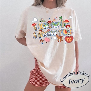 May include: An ivory t-shirt featuring a graphic print of Toy Story characters including Woody, Buzz Lightyear, Jessie, Rex, Bullseye, and Hamm. The text reads I KNEW IT I KNEW YOU. The design includes small stars, clouds, a rocket, and a red heart with TS.