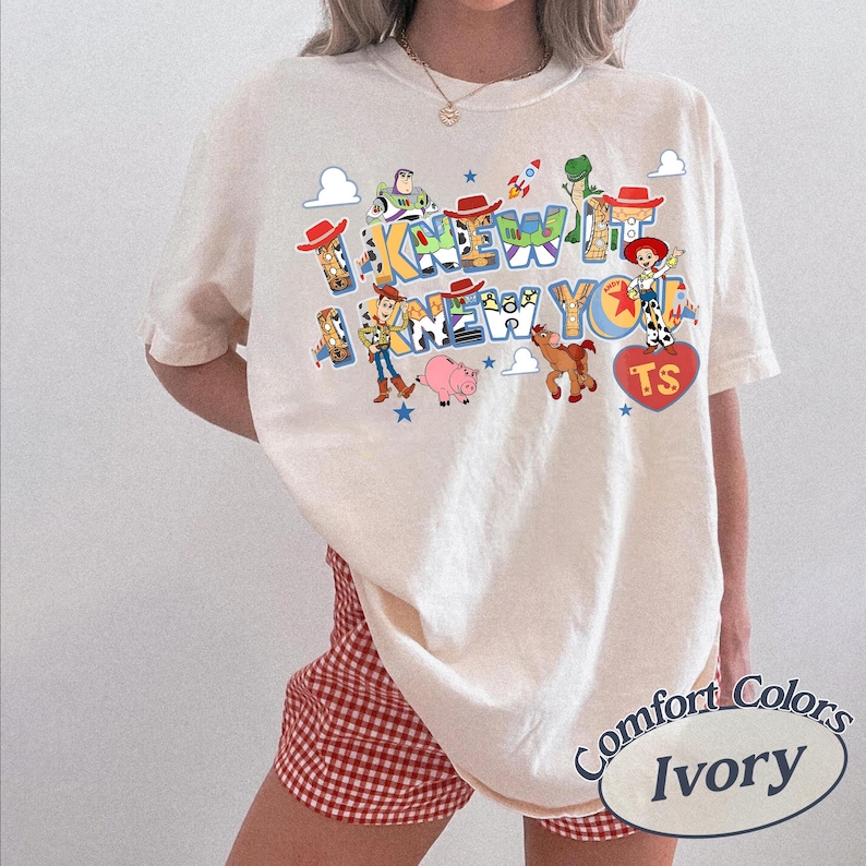 May include: An ivory t-shirt featuring a graphic print of Toy Story characters including Woody, Buzz Lightyear, Jessie, Rex, Bullseye, and Hamm. The text reads I KNEW IT I KNEW YOU. The design includes small stars, clouds, a rocket, and a red heart with TS.