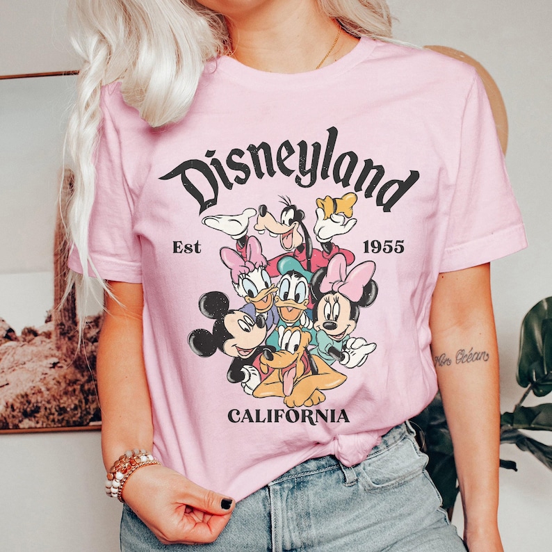 May include: A light pink t-shirt featuring the word "Disneyland" in a black, vintage font, with the text "Est 1955 California." The shirt displays a graphic of Disney characters including Mickey Mouse, Minnie Mouse, Donald Duck, and Goofy.