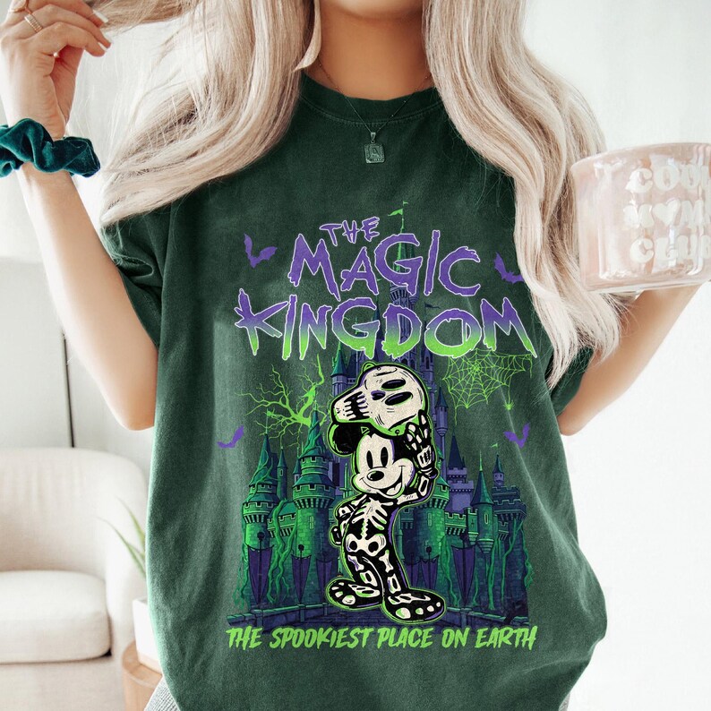 May include: Dark green t-shirt with a graphic featuring Mickey Mouse in a skeleton costume holding a mask. The text "THE MAGIC KINGDOM" and "THE SPOOKIEST PLACE ON EARTH" are printed in purple and green.
