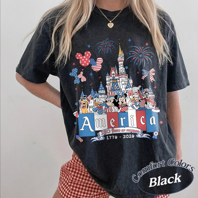 May include: A black acid-wash t-shirt featuring a graphic of a castle, fireworks, and cartoon characters in patriotic red, white, and blue. The text reads America, 250 Years of Freedom, and 1776-2026. The shirt has short sleeves and a crew neckline.