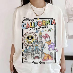 Puede incluir: Camiseta de color crema con un gr&aacute;fico colorido de Disney California Adventure. El dise&ntilde;o incluye a Mickey Mouse en una noria, Daisy Duck, un castillo y el texto "Disney California Adventure" y "Your Name".