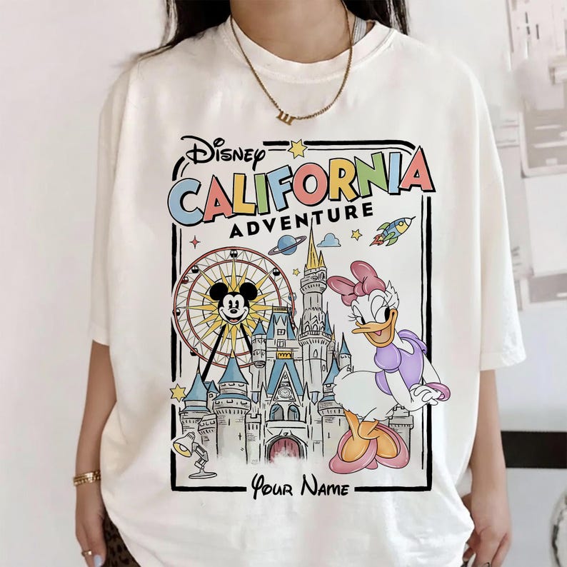 Puede incluir: Camiseta de color crema con un gr&aacute;fico colorido de Disney California Adventure. El dise&ntilde;o incluye a Mickey Mouse en una noria, Daisy Duck, un castillo y el texto "Disney California Adventure" y "Your Name".