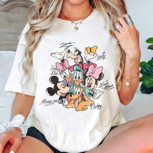 May include: Cream-colored t-shirt featuring a colorful graphic of Disney characters including Mickey Mouse, Minnie Mouse, Donald Duck, Daisy Duck, Goofy, and Pluto. The characters' names are written above or below them in a handwritten style.