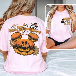 May include: Light pink t-shirt featuring a Halloween-themed graphic. The design includes a smiling jack-o'-lantern with Mickey Mouse ears, spiderwebs, and the text "Walt Disney World EST. 1971". A smaller version of the design is on the front pocket.