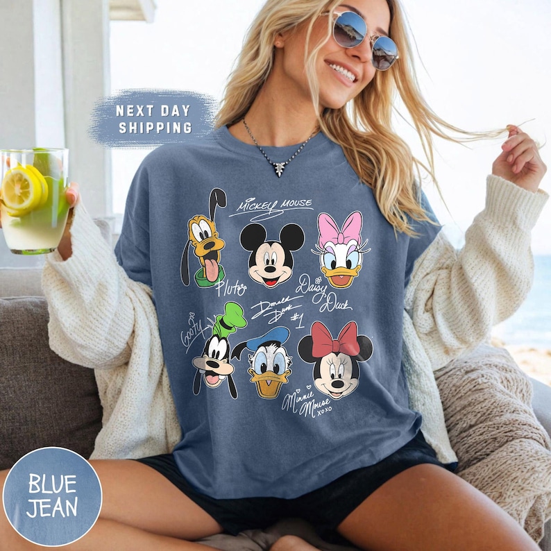 May include: A blue jean t-shirt with a graphic print of Mickey Mouse and his friends. The shirt features the characters Mickey Mouse, Minnie Mouse, Donald Duck, Daisy Duck, Goofy, and Pluto. Each character has their signature under their image.