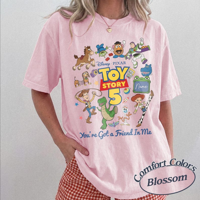 May include: A light pink t-shirt featuring a colorful graphic of Disney Pixar Toy Story characters including Woody, Buzz Lightyear, Jessie, Rex, and Forky. The design includes the text Toy Story 5 and You're Got a Friend In Me, with a blank space for a name.
