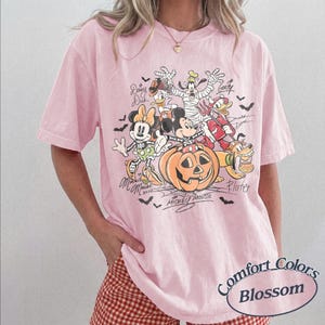 May include: A light pink t-shirt featuring a graphic print of Disney characters including Mickey Mouse, Minnie Mouse, Donald Duck, Daisy Duck, Goofy, and Pluto dressed in Halloween costumes around a large orange jack-o'-lantern with bats.