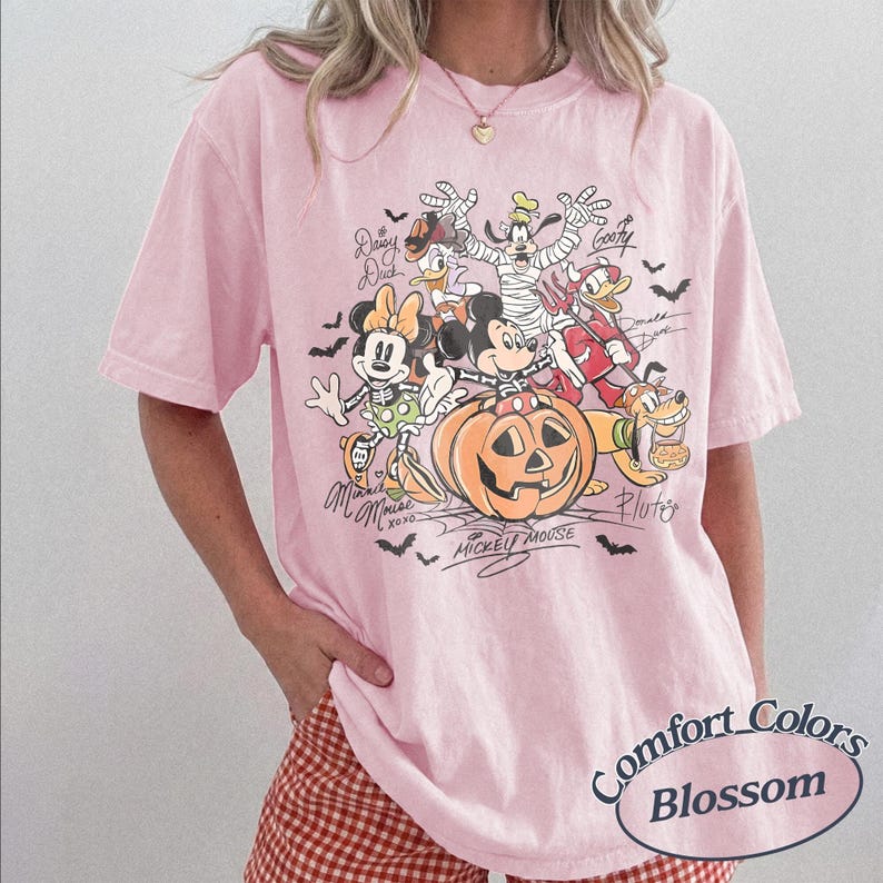 May include: A light pink t-shirt featuring a graphic print of Disney characters including Mickey Mouse, Minnie Mouse, Donald Duck, Daisy Duck, Goofy, and Pluto dressed in Halloween costumes around a large orange jack-o'-lantern with bats.