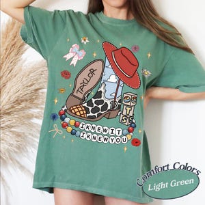 May include: A light green t-shirt featuring a graphic print of a cow-print cowboy boot with a red hat, the name Taylor, the number 13, and blocks spelling BTS. Below, a beaded necklace graphic displays the text I KNEW IT I KNEW YOU.