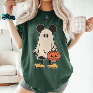 May include: A green t-shirt with a white ghost wearing Mickey Mouse ears and holding a jack-o'-lantern. The ghost is wearing black shoes.