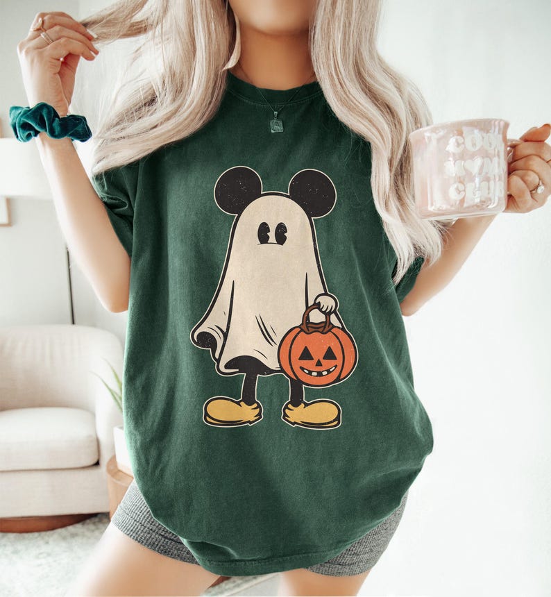 May include: A green t-shirt with a white ghost wearing Mickey Mouse ears and holding a jack-o'-lantern. The ghost is wearing black shoes.