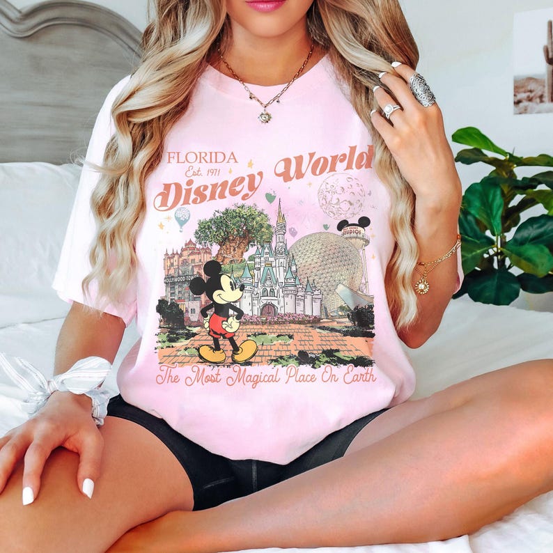 May include: A light pink t-shirt featuring a graphic of Mickey Mouse in front of Cinderella's castle and Spaceship Earth at Disney World. The text "Florida Disney World" and "The Most Magical Place On Earth" are also printed on the shirt.