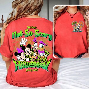 Op de afbeelding: Rood T-shirt met een Halloween-ontwerp met Mickey Mouse, Minnie Mouse, Goofy, Donald Duck en Daisy Duck. De achterkant van het shirt heeft de tekst "Mickey's Not-So-Scary Halloween Party 2025". De voorkant heeft een kleiner ontwerp met een skeletkarakter.