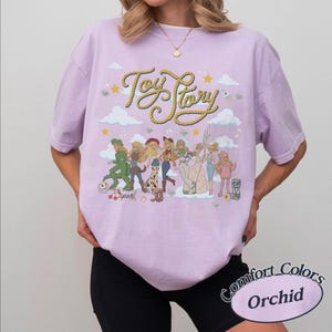 May include: A light purple short-sleeve t-shirt featuring a graphic print of Toy Story characters in various outfits. The text Toy Story is written in a yellow rope-style font above the characters, surrounded by clouds, stars, and musical notes.