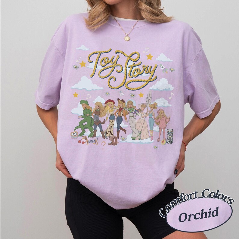 May include: A light purple short-sleeve t-shirt featuring a graphic print of Toy Story characters in various outfits. The text Toy Story is written in a yellow rope-style font above the characters, surrounded by clouds, stars, and musical notes.
