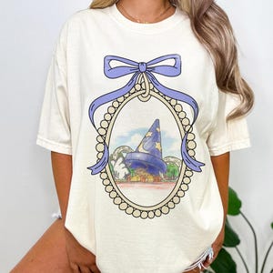 May include: A white t-shirt with a purple bow and a framed image of a blue and white hat with stars. The hat is on top of a building with a Mickey Mouse hand reaching out.