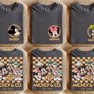 May include: Three gray t-shirts with a checkered pattern and Mickey Mouse characters. The shirts have the text "Mickey & Co. Est. 1928" printed on the bottom. The shirts are labeled "Daddy", "Mommy", and "Custom".