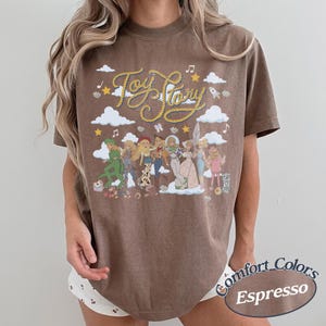 May include: A brown short-sleeve t-shirt featuring a graphic print with the text Toy Story in yellow rope-style lettering. The design includes white clouds, yellow stars, and a group of illustrated characters from the Toy Story film franchise.
