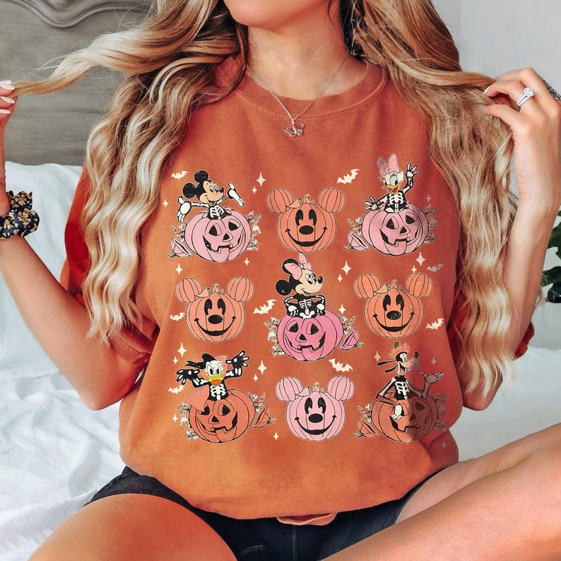 May include: Orange-colored t-shirt featuring cartoon characters on Halloween-themed pumpkins. The design includes Mickey Mouse, Minnie Mouse, Donald Duck, and Goofy, with bats and stars. The pumpkins are pink and orange.