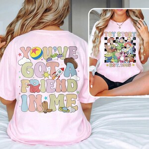 May include: Pink t-shirt with a colorful graphic that says "You've got a friend in me" and features characters from the Toy Story movie franchise. The back of the shirt has a black and white checkered graphic with the text "Toy Story Est. 1995".