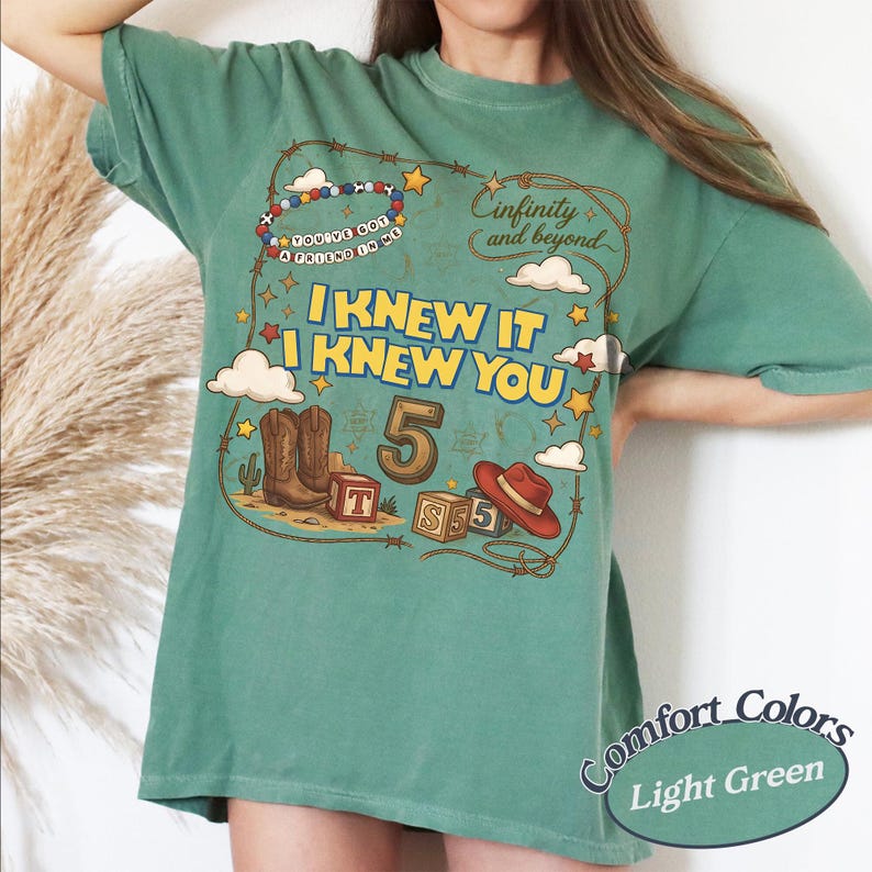 May include: A light green t-shirt featuring a graphic print with the text "I KNEW IT I KNEW YOU", "Infinity and beyond", and "YOU'VE GOT A FRIEND IN ME". The design includes cowboy boots, a red hat, alphabet blocks, stars, and clouds.