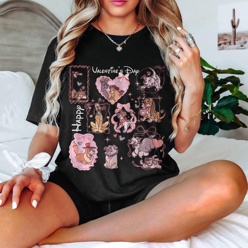 May include: Black t-shirt featuring various dog illustrations in pink frames and heart shapes. The shirt has the words "Happy" and "Valentine's Day" printed on it. The design includes cartoon dogs with bows and other decorative elements.