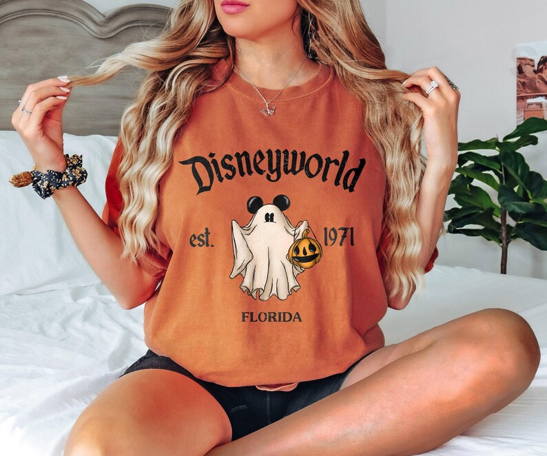 Op de afbeelding: Oranje T-shirt met een zwarte afbeelding van een geest die een pompoen vasthoudt. De afbeelding zegt "Disneyworld est. 1971 Florida".