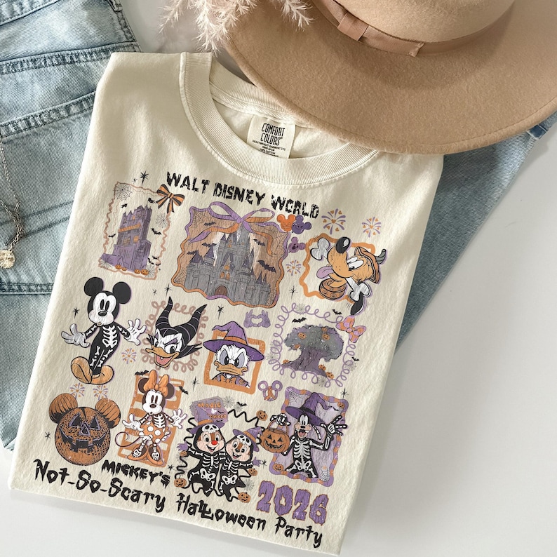 May include: Cream-colored t-shirt with a colorful graphic featuring Disney characters in Halloween costumes. The shirt includes the text "WALT DISNEY WORLD" and "MICKEY'S Not-So-Scary Halloween Party 2026". A tan hat and blue jeans are in the background.
