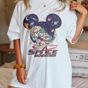 May include: White t-shirt with a graphic of Mickey Mouse's head with a space scene inside. The graphic includes Mickey Mouse, Minnie Mouse, and Goofy riding a space mountain ride. The text "Space Mountain" is printed below the graphic.