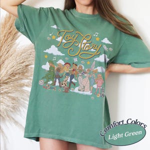 May include: A sage green short-sleeve t-shirt featuring a graphic print with the text Toy Story in yellow rope-style lettering. The design includes illustrations of various characters, white clouds, yellow stars, and musical notes across the front.