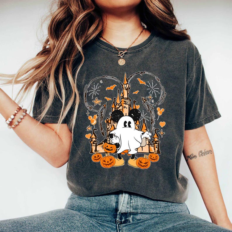 May include: Dark grey t-shirt with a Halloween graphic. The design features a cartoon ghost, pumpkins, a castle, and fireworks. The graphic is in orange, black, and white.