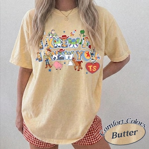 May include: A pale yellow short-sleeve t-shirt featuring a graphic print of various animated characters and the text I KNEW IT I KNEW YOU. The design includes colorful illustrations of a space ranger, a cowboy, a pig, a horse, and a red heart with TS.