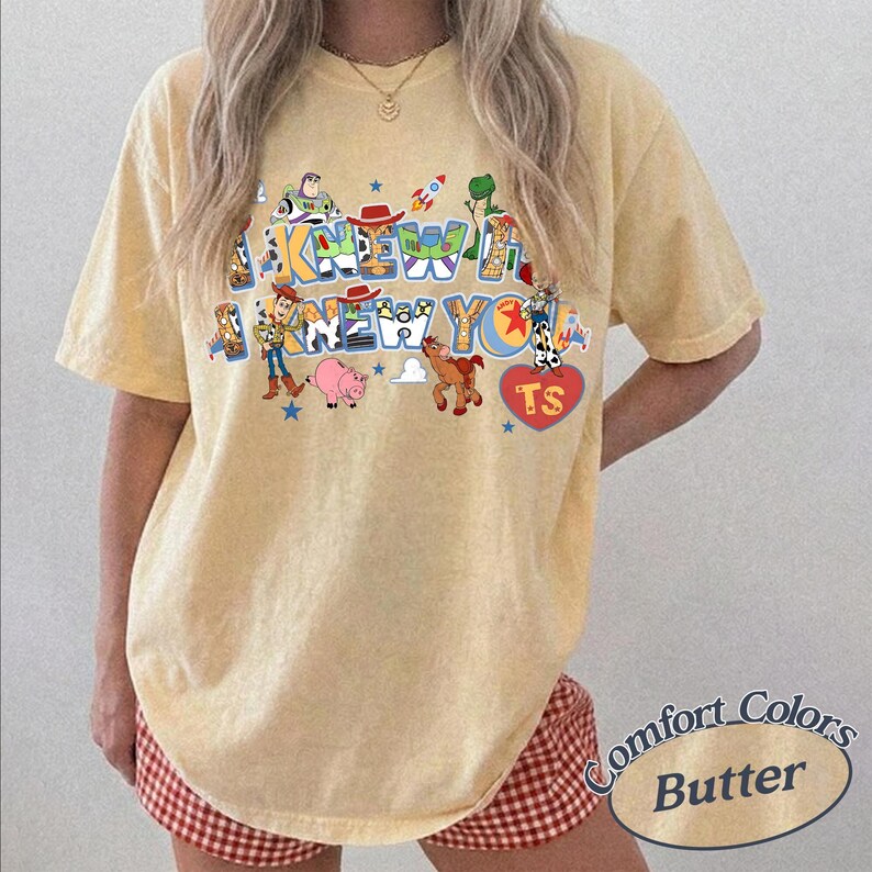 May include: A pale yellow short-sleeve t-shirt featuring a graphic print of various animated characters and the text I KNEW IT I KNEW YOU. The design includes colorful illustrations of a space ranger, a cowboy, a pig, a horse, and a red heart with TS.