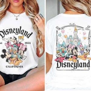 May include: White t-shirt with a graphic print of Mickey Mouse and friends in front of Disneyland. The text "Disneyland Est 1955 California" is printed on the front of the shirt. The back of the shirt features a graphic print of Disneyland castle and Mickey Mouse and friends. The text "Disneyland 1955" is printed on the back of the shirt.