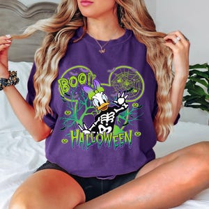 May include: Purple t-shirt featuring a Halloween-themed Daisy Duck design. The graphic includes a skeleton, spiderwebs, and the words "BOO!" and "HALLOWEEN" in green. The design is set within a Mickey Mouse ear outline.