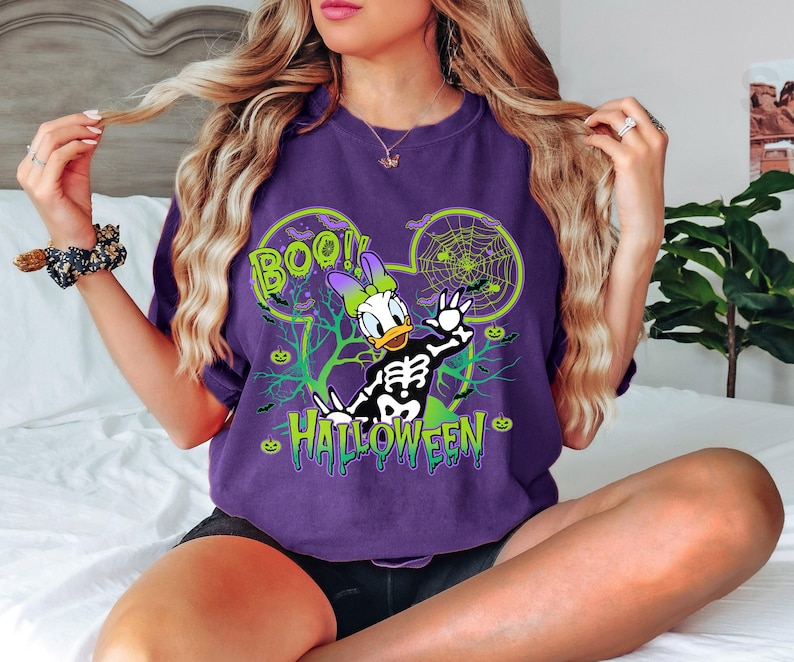 May include: Purple t-shirt featuring a Halloween-themed Daisy Duck design. The graphic includes a skeleton, spiderwebs, and the words "BOO!" and "HALLOWEEN" in green. The design is set within a Mickey Mouse ear outline.