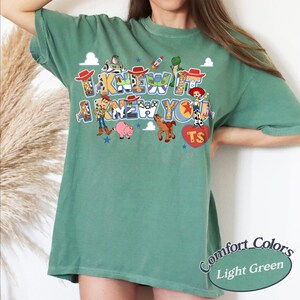 May include: A light green short-sleeve t-shirt featuring a graphic print of Toy Story characters including Woody, Buzz Lightyear, Jessie, Rex, Hamm, and Bullseye. The text reads I KNEW IT I KNEW YOU with a red heart containing the letters TS.